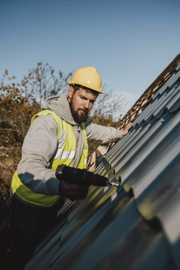 A construction worker in a yellow hard hat and safety vest uses a power drill to install metal roofing panels on a sloped roof, ensuring durability and safety for Emergency Roof Repair in Boca Raton.