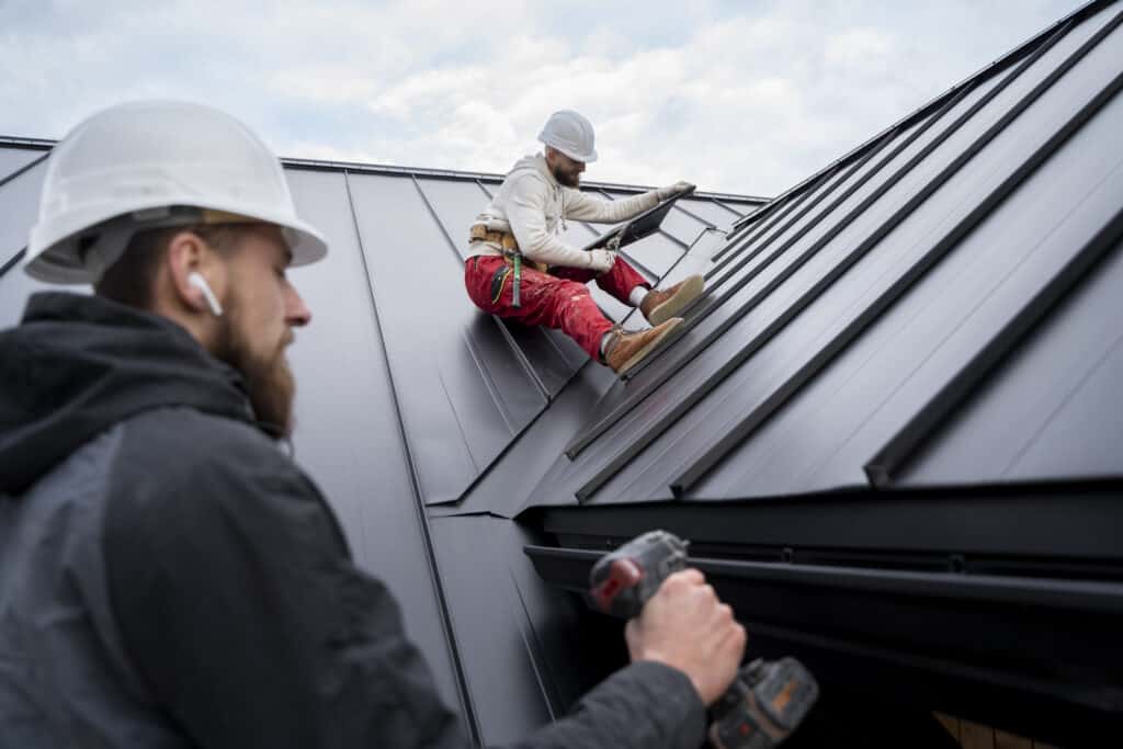 Roof Leak Repair Cost - Fix that leak! 1 Two workers in safety gear install metal roofing panels during a local roof inspection; one uses a drill while the other adjusts a panel on the sloped roof.