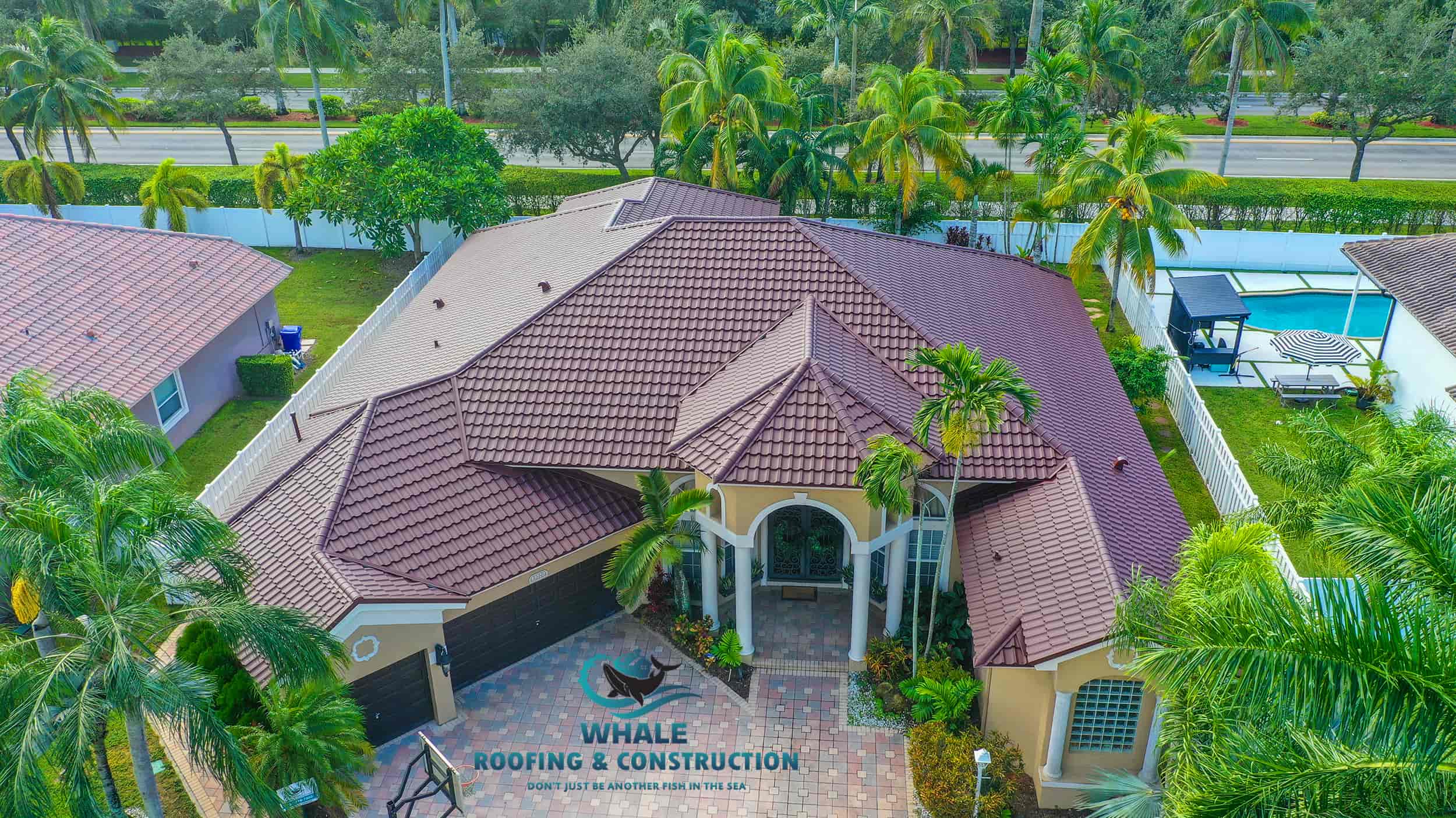Aerial view of a single-story house with a brown tiled roof, surrounded by palm trees and a paved driveway, with "Whale Roofing & Construction" logo.