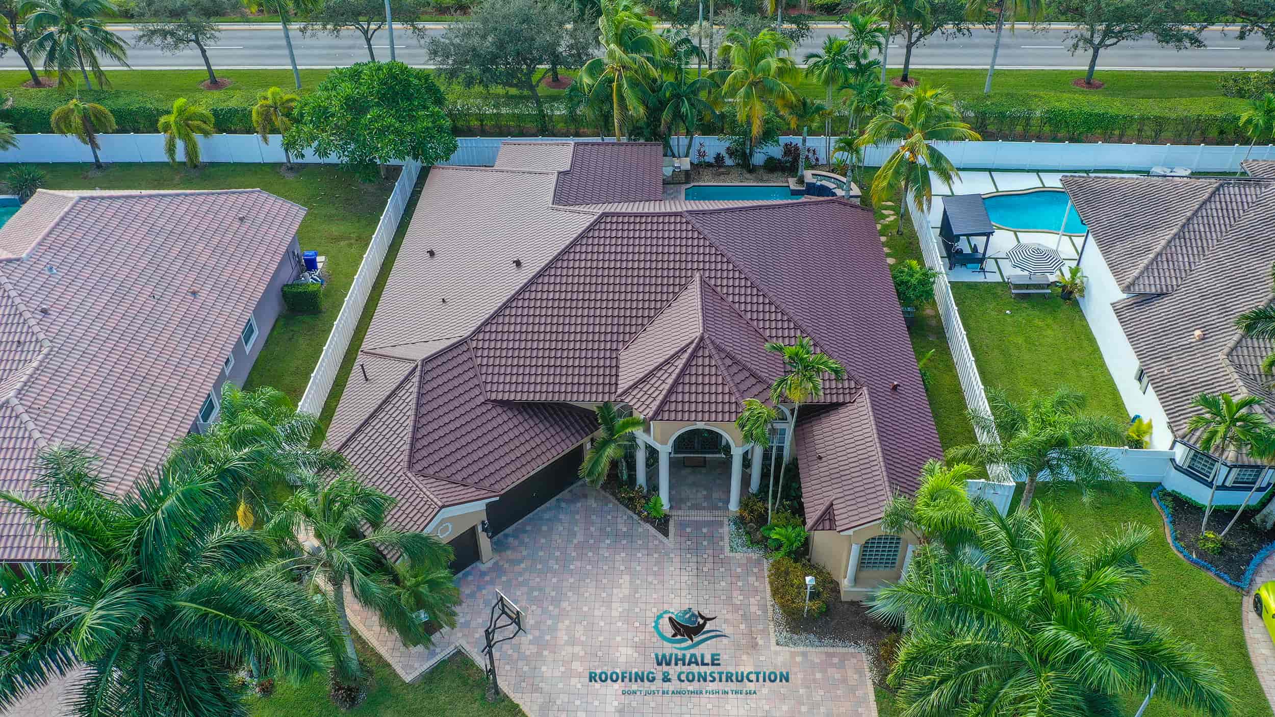 Aerial view of a single-story house with a brown tiled roof, a paved driveway, pool in the backyard, and manicured lawn, surrounded by neighboring homes and trees.