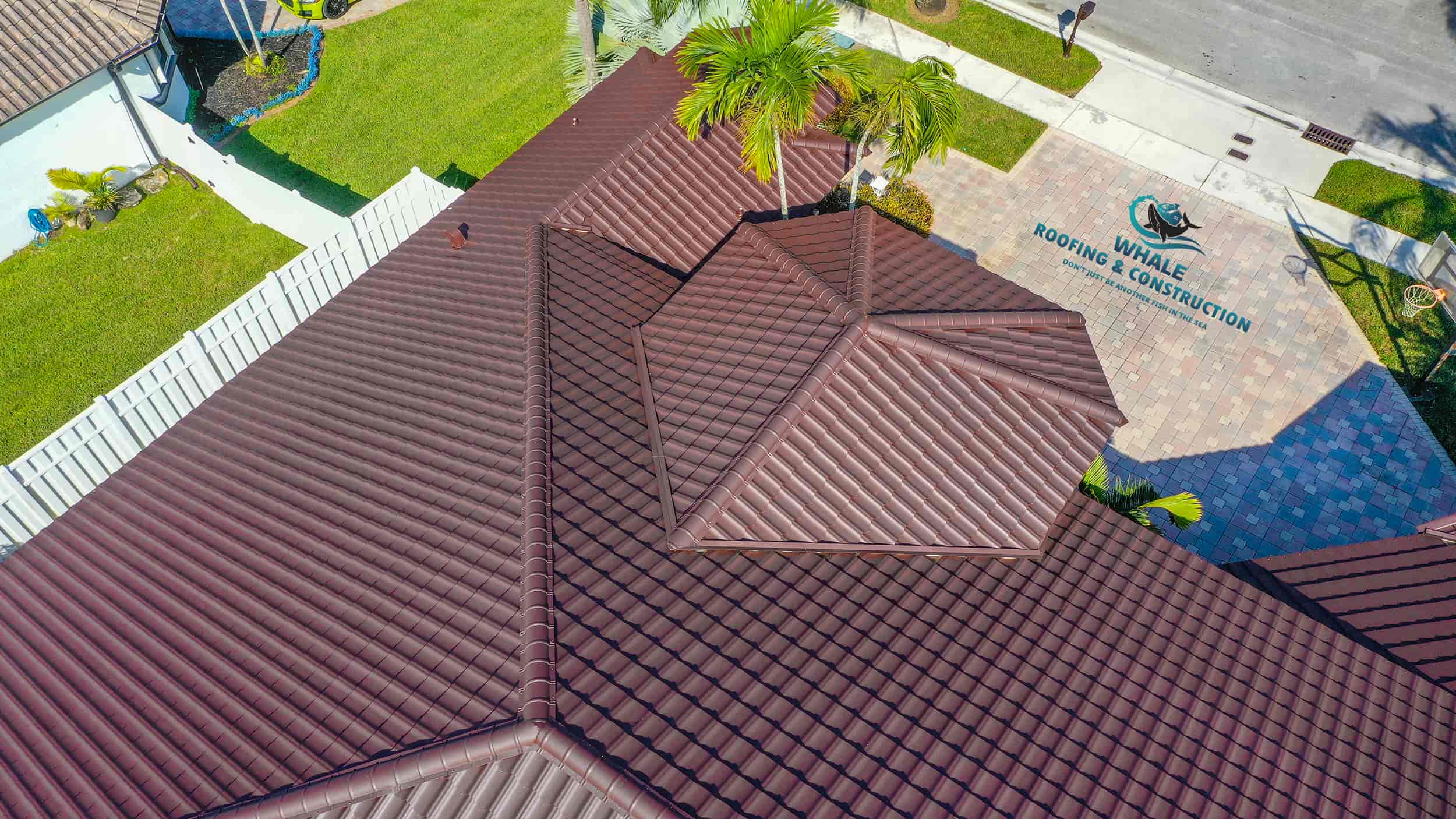 Aerial view of a brown tile roof on a residential home with a driveway displaying "Whale Roofing & Construction" logo and palm trees nearby.