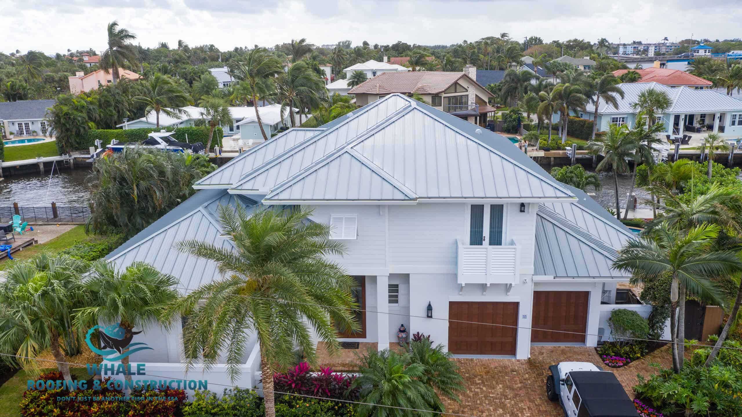 Aerial view of a two-story house with a metal roof, surrounded by palm trees, a canal, and neighboring homes; Hale Roofing & Construction logo is visible, highlighting expert roofing services Boca Raton residents can trust.