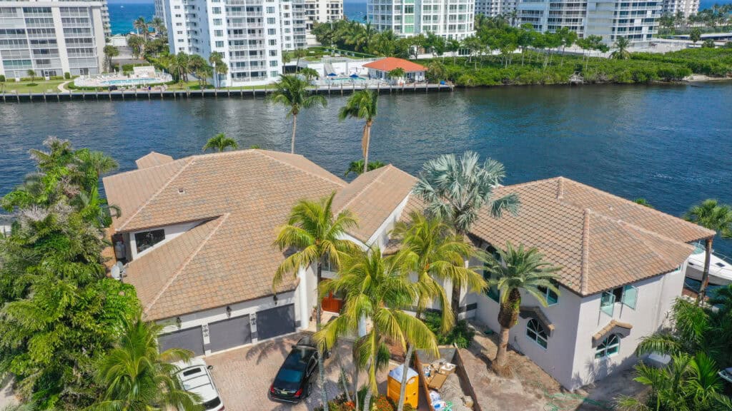 A large waterfront house with a tile roof by one of the best roofers in South Florida is surrounded by palm trees, adjacent to a canal, with high-rise buildings visible across the water.