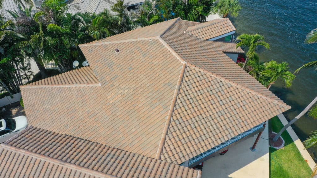 Aerial view of a beige tile roof on a house surrounded by palm trees, with water visible nearby—a perfect example of the quality work offered by commercial roofing contractors in South Florida.