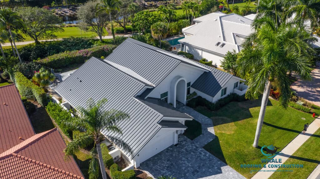 Aerial view of a white house with a dark gray metal roof—perfect for protecting against storm hail damage—surrounded by palm trees, a paved driveway, and neighboring homes.