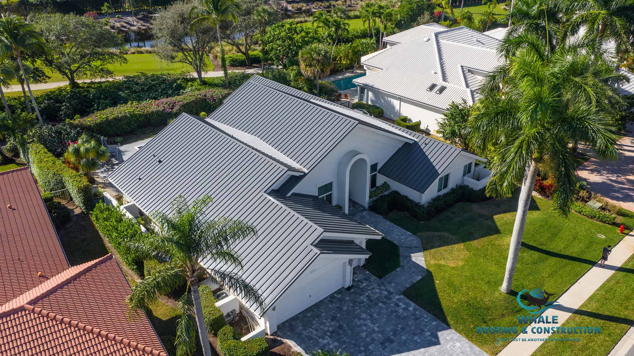 Aerial view of a white house with a dark gray metal roof—perfect for protecting against storm hail damage—surrounded by palm trees, a paved driveway, and neighboring homes.