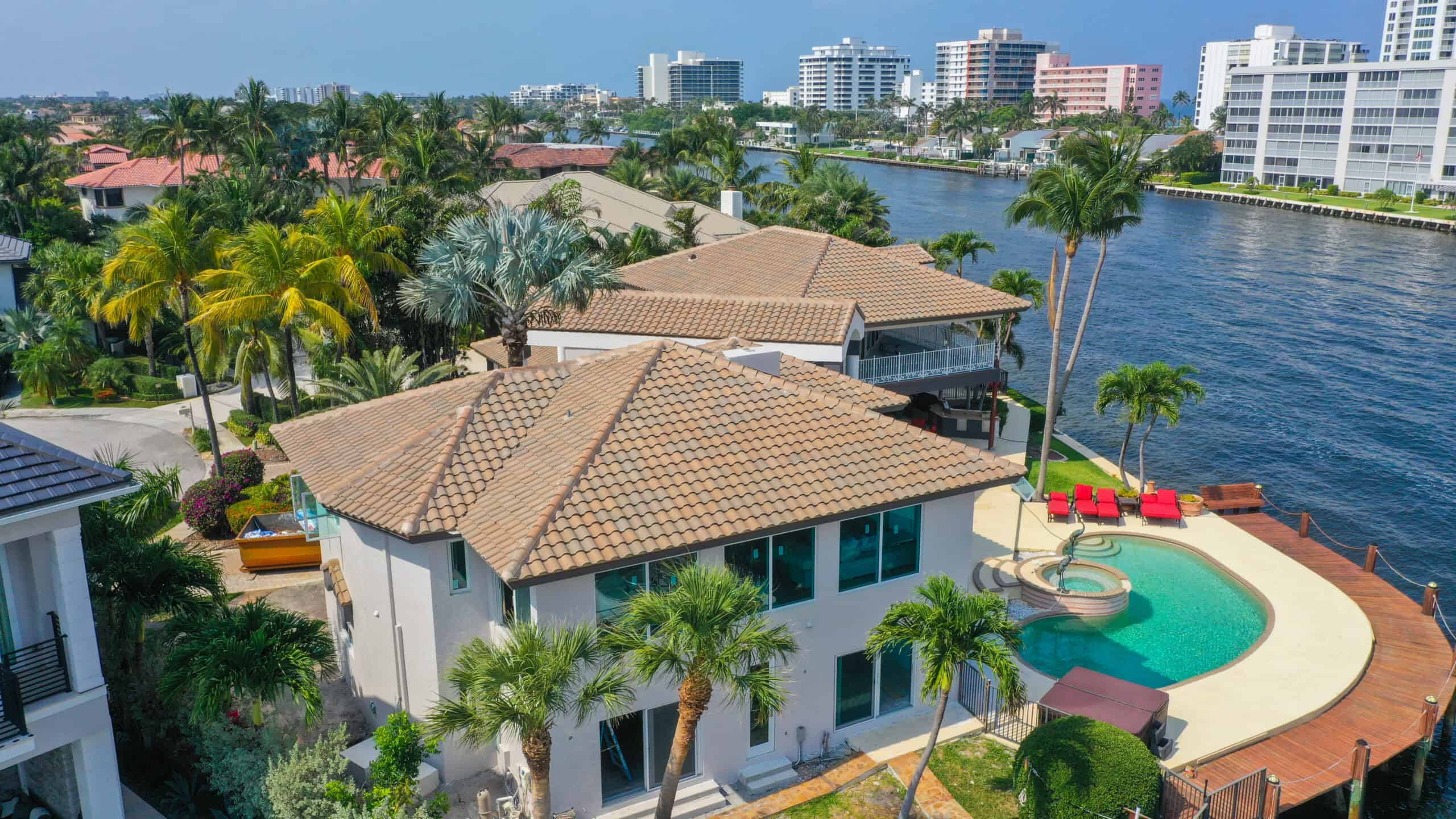 A waterfront house with a tiled roof—expertly maintained with Roofing Consultations—features a private pool and patio overlooking a canal lined with palm trees and city buildings.
