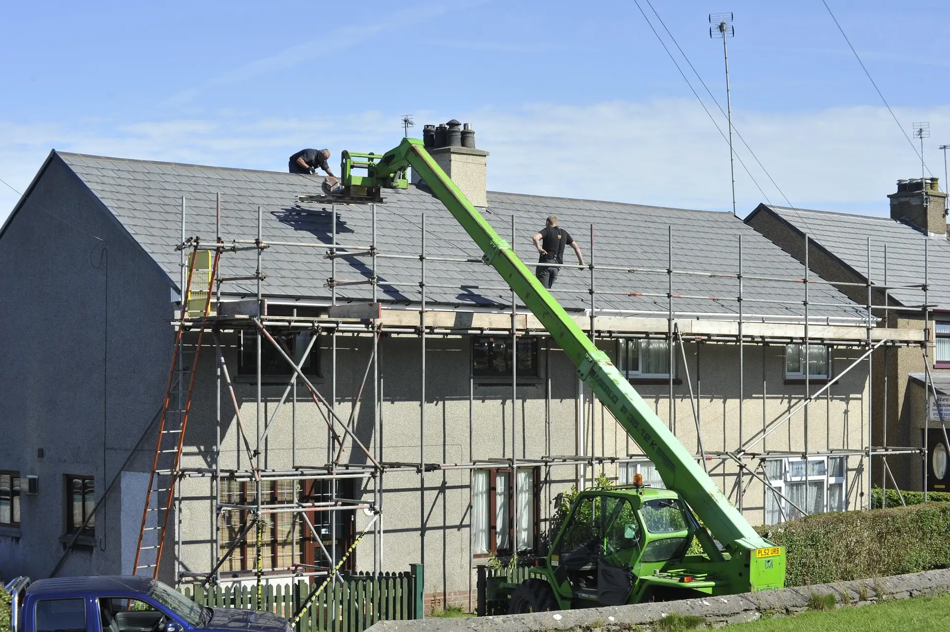 Two workers repair the roof of a house using scaffolding and a green telescopic handler on a sunny day.