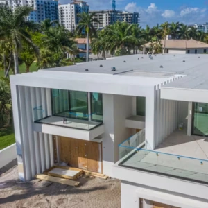 Whale Roofing Services 5 Two-story modern house under construction with large glass windows and balconies, set in a tropical area with palm trees and high-rise buildings in the background—perfect for quality roof installation Boca Raton services.
