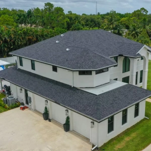 Whale Roofing Services 4 Aerial view of a two-story modern house with a dark gray roof, light exterior walls, three garage doors, and a driveway surrounded by green lawn and trees—ideal for those seeking expert roofing services or roof installation in Boca Raton.
