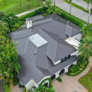 Whale Roofing Services 2 Aerial view of a large house with a dark gray metal roof, surrounded by palm trees and greenery, with Whale Roofing & Construction logo in the bottom right corner.