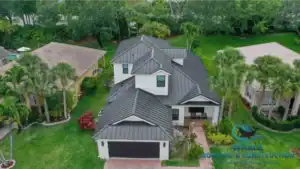 Aerial view of a modern two-story house with a dark metal roof, white exterior walls, and a brick driveway, surrounded by green lawns and palm trees.