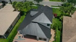 Aerial view of a two-story house with a black metal roof, surrounded by green lawns and neighboring homes, with a street visible at the top.