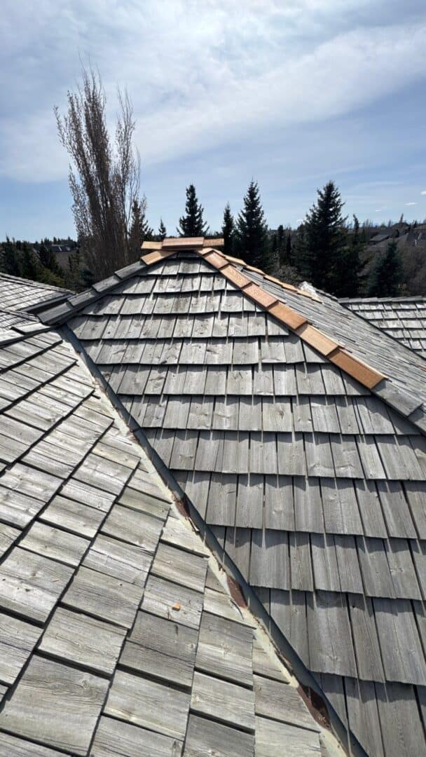 A cedar shake roof in Calgary showing weathered wood shingles with recent ridge cap repairs, highlighting the aging process and maintenance challenges common to wood roofing.