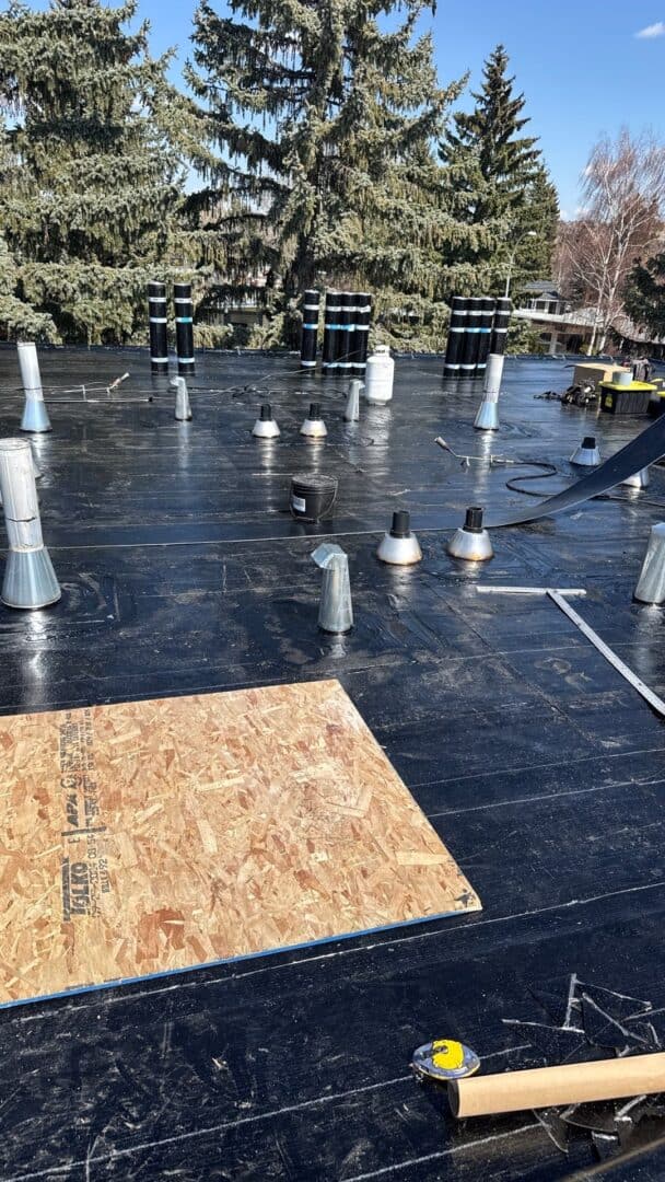 Flat roof installation in progress with new high-dome goose necks and furnace vents properly sealed and set in place on a fresh torch-on membrane. Roofing tools and materials are visible across the work area.