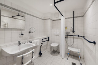 Behindertengerechtes Bad: Schwellenlose, befahrbare Dusche mit Handlauf und Duschhocker, unterfahrbares Waschbecken mit Waschhocker und verstellbarem Wand- und Kosmetikspiegel. WC mit seitlichem Stützgriff