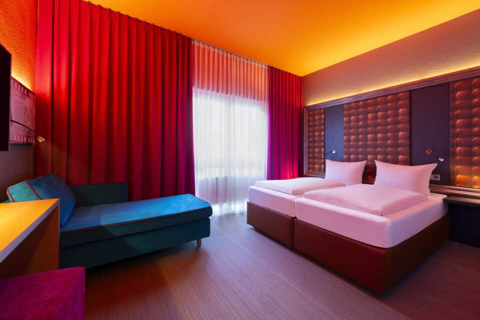 Behindertengerechtes Zimmer – zwei elektrisch verstellbare Einzelbetten und blauem Sofa. Regal als Nachttisch zwischen den Betten und wandmontierte, schwenkbare Lampen.