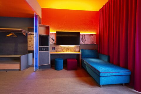 Behindertengerechtes Zimmer – Arbeitsbereich und Garderobe. Offene Garderobe links, unterfahrbarer Schreibtisch mit TV darüber, Minibar und Safe in Regal mit Stauraum, im Sitzen erreichbar, zwei Hocker