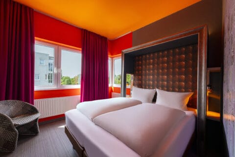 Doppelzimmer – Blickrichtung, Bett und Fenster. Doppelbett durchgehend bezogen, Nachttische, Fenster, Kunststoff-Flechtsessel
