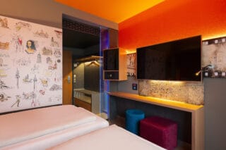 Einzelzimmer – Blick vom Bett. Arbeitsplatz, TV, Safe, Minibar, offene Garderobe, Sessel mit Fellauflage, zwei Hocker