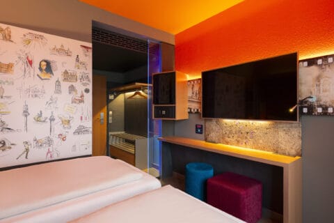 Einzelzimmer – Blick vom Bett. Arbeitsplatz, TV, Safe, Minibar, offene Garderobe, Sessel mit Fellauflage, zwei Hocker