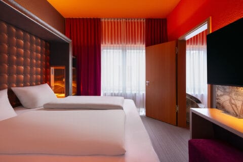 Familienzimmer – Blick zur offenen Verbindungstür. Doppelbett mit Poster-Kopfteil links, Arbeitsplatz rechts, Sessel vor Verbindungstür