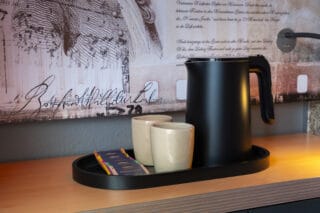 Junior-Suite – Teestation: Wasserkocher mit zwei Keramiktassen und Teebeuteln auf Tablett, dahinter Motivtapete mit Texten in historisch anmutender Schrift. Unten zweitürige Kommode.