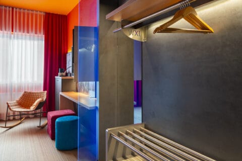 Junior-Suite – Perspektive vom Eingang. Offene Garderobe mit Kleiderstange rechts, Arbeitsplatz mit Hockern mittig, Schaukelstuhl am Fenster links