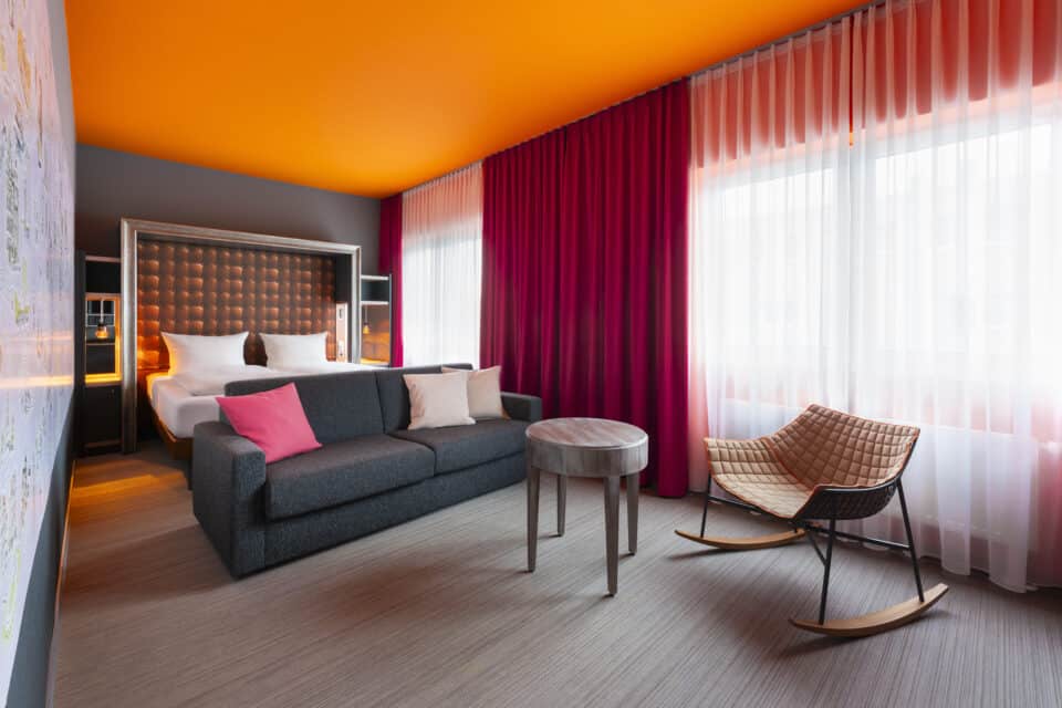 Junior-Suite – Perspektive vom Wohnbereich zum Bett. Runder Beistelltisch, Sofa mit Kissen, Doppelbett mit gerahmtem Kopfteil, Hannover-Motivwand links, Fenster rechts.