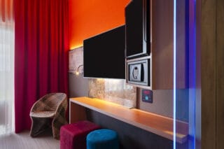 Zimmer – Blick zum geflochtenen Sessel und Arbeitsplatz. Schreibtisch, TV/Safe/Minibar in Augenhöhe, Kommode, zwei Hocker
