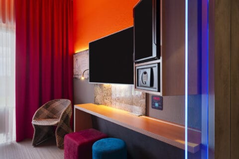 Zimmer – Blick zum geflochtenen Sessel und Arbeitsplatz. Schreibtisch, TV/Safe/Minibar in Augenhöhe, Kommode, zwei Hocker