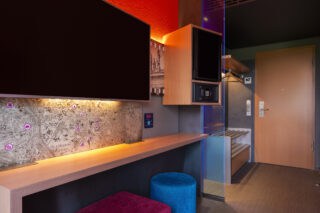 Zimmer – Blick zu Eingang und Arbeitsplatz. Zimmertür, Garderobe, Schreibtisch, TV/Safe/Minibar in Augenhöhe, Kommode