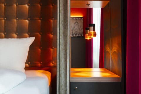 Zimmer - Detailansicht vom Bett, rechts Nachttisch mit dunkler Schublade, leuchtende Lampe