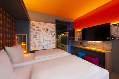 Zweibettzimmer– Blick vom Bett. Arbeitsplatz, TV, Safe, Minibar, offene Garderobe, Sessel mit Fellauflage, zwei Hocker