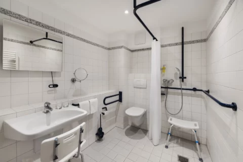 Behindertengerechtes Bad: Schwellenlose, befahrbare Dusche mit Handlauf und Duschhocker, unterfahrbares Waschbecken mit Waschhocker und verstellbarem Wand- und Kosmetikspiegel. WC mit seitlichem Stützgriff