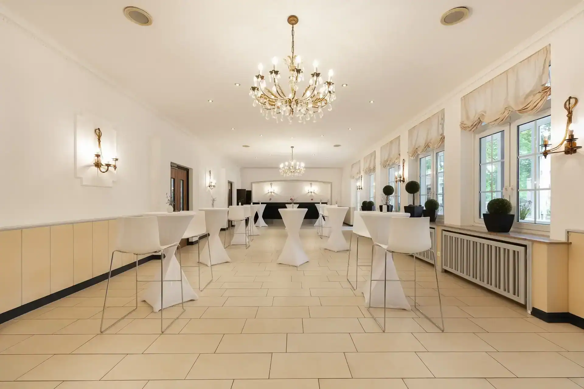 Tagungsraum IX (Bistro) – Frontalperspektive. Stehtische mit Barhockern in Reihe, zwei Kronleuchter, Wandleuchten an allen Wänden, Fensterfront rechts mit Vorhängen und Pflanzendekoration, Fliesenboden