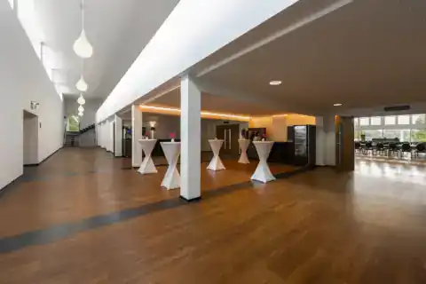 CongressCentrum Foyer – Fünf Stehtische mit Hussen, Getränkestation, langer Flur mit Treppe links, offener Durchgang zum Saal rechts, Holzboden, Oberlicht und Pendelleuchten