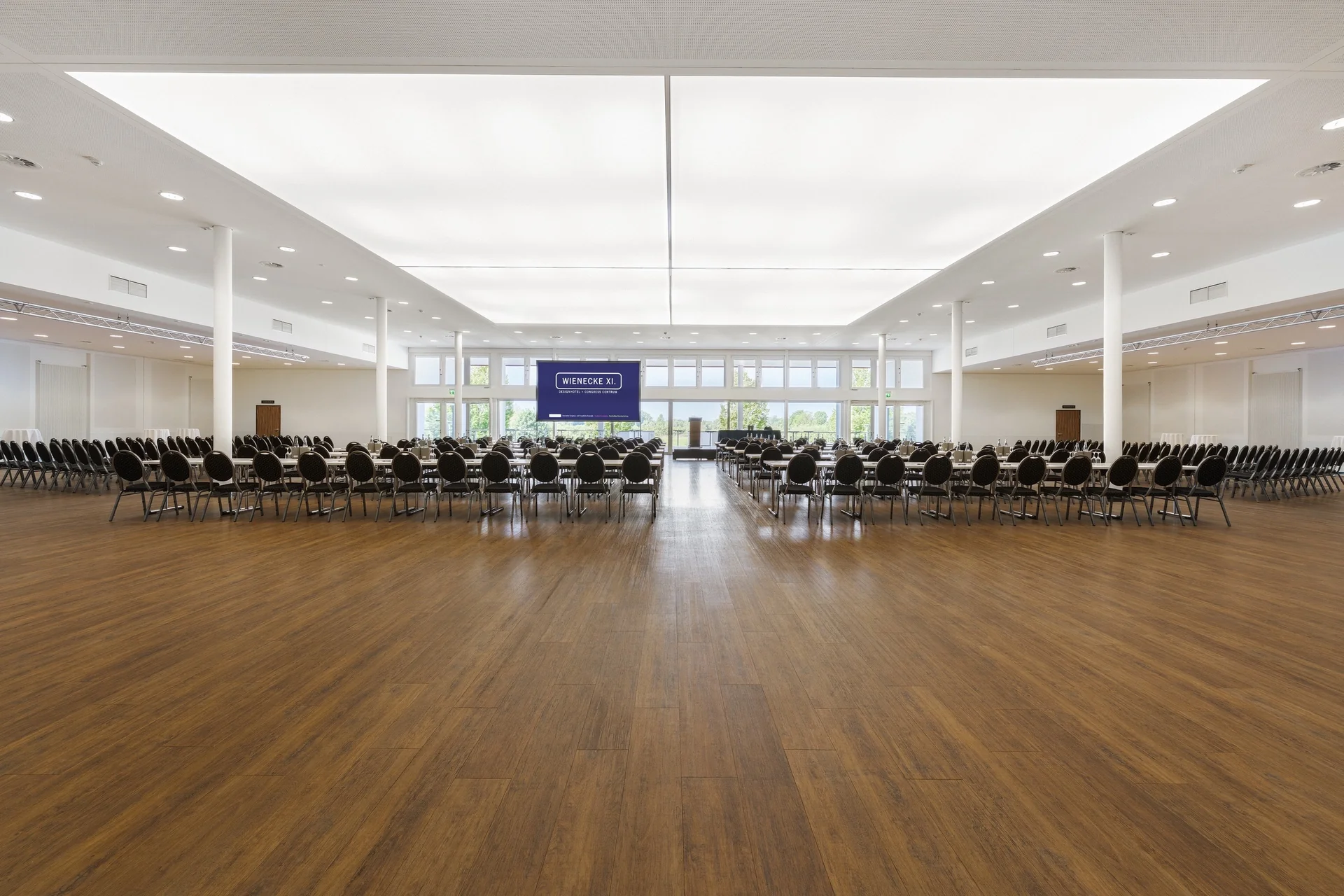CongressCentrum – Frontalperspektive. Parlamentarische- und Reihenbestuhlung, LED-Display mittig vor Fensterfront zum Landschaftsschutzgebiet, großes Oberlicht, Säulen, Holzboden