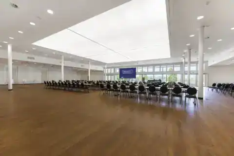 CongressCentrum - Diagonal perspective. Row seating with tables, three pillars on the left and right, LED display in front of window front with view of the landscape conservation area behind, bar tables on the left, large illuminated skylight, wooden floor