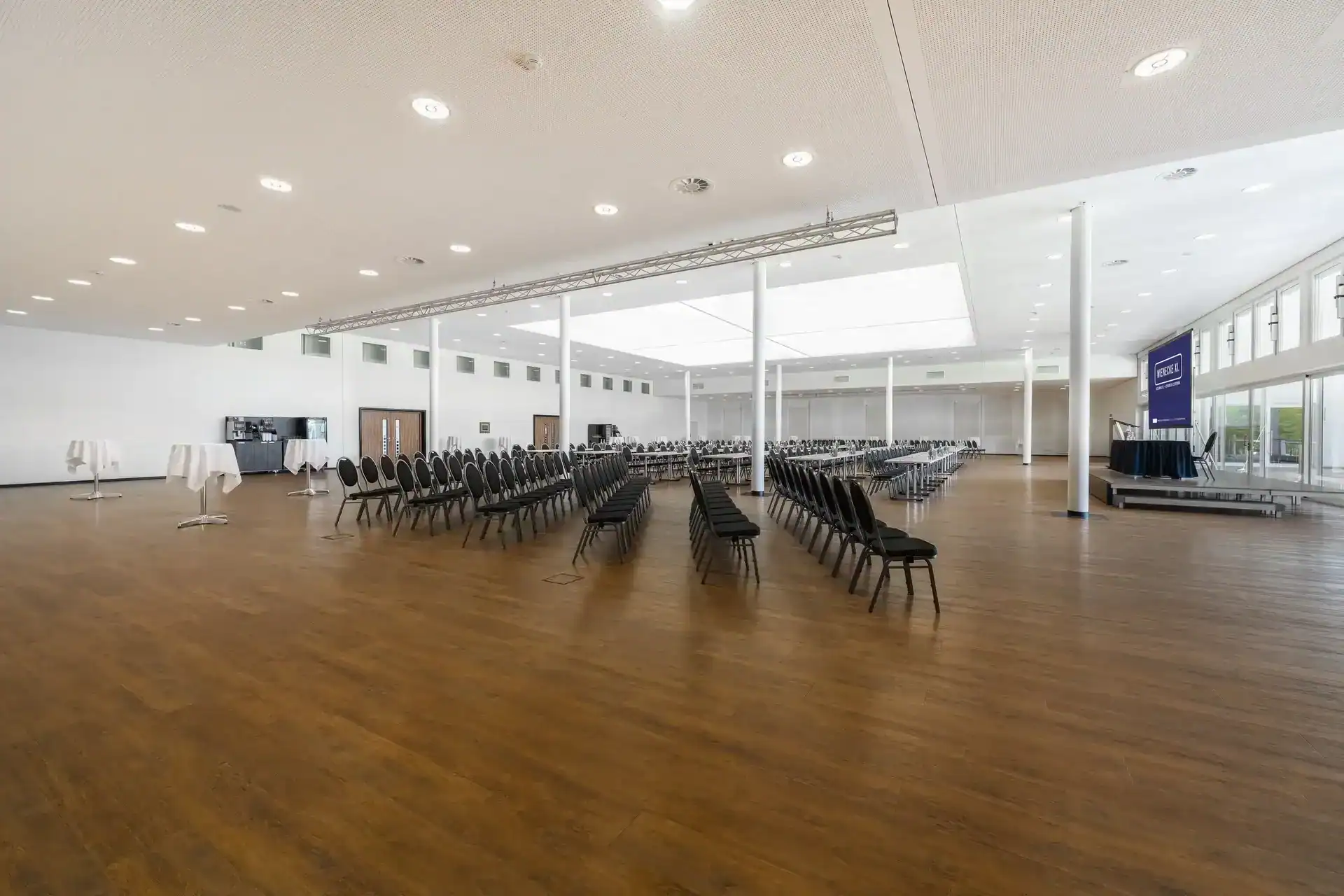 CongressCentrum – Seitliche Rückansicht. Reihenbestuhlung, Bühne mit LED-Display rechts, Fensterfront mit Terrasse dahinter, drei Stehtische links, großes Oberlicht, Traversensystem, Holzboden