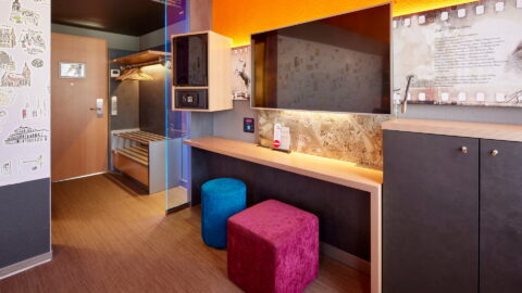 Doppelzimmer – Blick zu Eingang und Arbeitsplatz. Zimmertür, Garderobe, Schreibtisch, TV/Safe/Minibar in Augenhöhe, Kommode