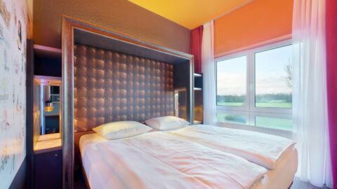 Doppelzimmer – Blickrichtung diagonal zu Bett und bodentiefem Fenster mit Vorhängen. Einzelbetten durchgehend bezogen, Nachttische