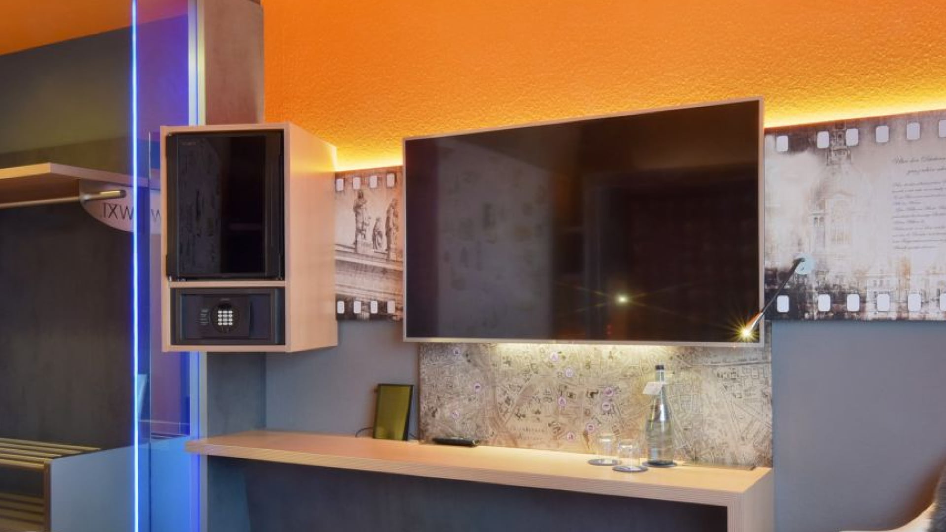 Einzelzimmer – Arbeitsplatzwand. Schreibtisch, v.l.n.r.: TV, Safe, Minibar in Augenhöhe, offener Schrank mit Garderobe