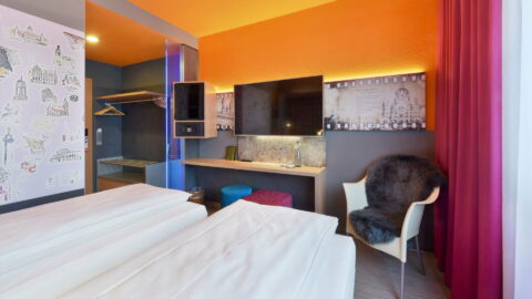 Einzelzimmer – Blick vom Bett. Arbeitsplatz, TV, Safe, Minibar, offene Garderobe, Sessel mit Fellauflage, zwei Hocker, Fenster