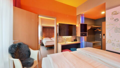 Familienzimmer – Blick vom Bett in beide Zimmer. Sessel mit Fellauflage, Arbeitsplatz mit TV/Minibar/Safe in Augenhöhe, offene Garderobe im Eingangsbereich, Verbindungstür zum zweiten Doppelbett