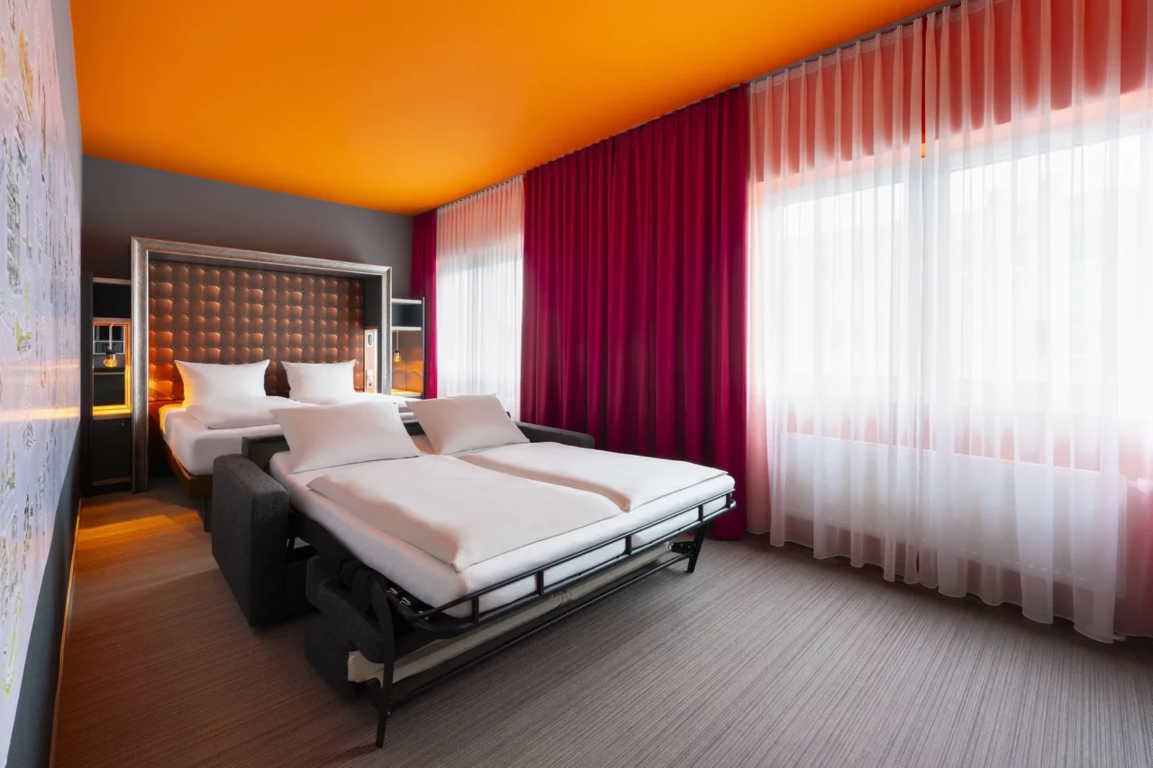 Familienzimmer – Perspektive über beide Schlafbereiche. Ausgeklapptes Schlafsofa vorne, Doppelbett mit umrahmtem Kopfteil hinten, Hannover-Motivwand, Fensterfront mit Vorhängen