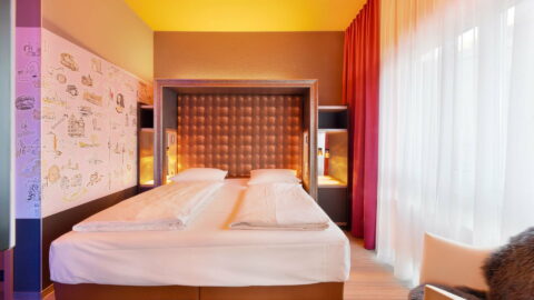 Familienzimmer – Doppelbett mit gerahmtem Kopfteil (Poster in Leder-Optik), daneben eingelassene Nachttische, Hannover-Motivwand, Fenster