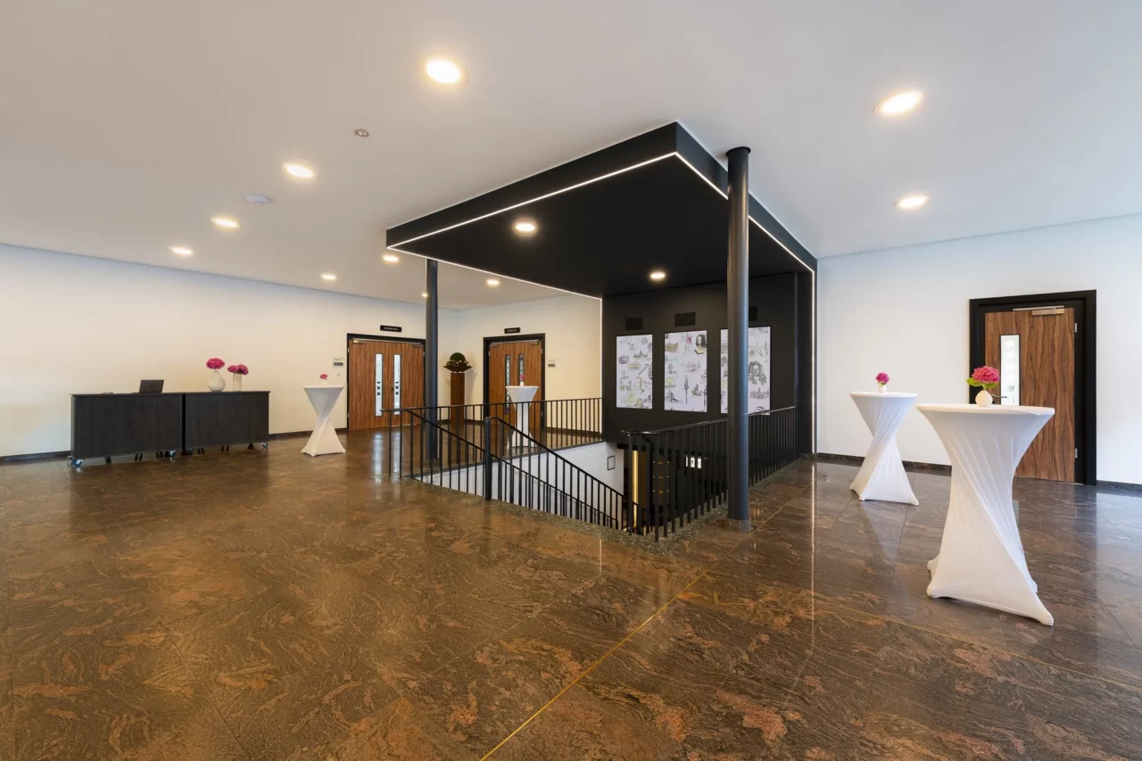 Ansicht vom Foyer des Kleinen Saals mit zwei weißen Stehtischen neben der Treppe, links zwei fahrbare dunkle Tische mit Blumen und Laptop, dunkle marmorierte Fliesen, helle Deckenspots, absteigende Treppe, drei LED-Displays über der Treppe mit Motiv-Tapete von Hannover