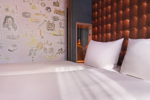 Junior-Suite – Nahaufnahme Kopfende Doppelbett: Decken, zwei Kissen, Poster-Kopfteil in Leder-Optik mit Nachttisch und Beleuchtung, Hannover-Motivwand links