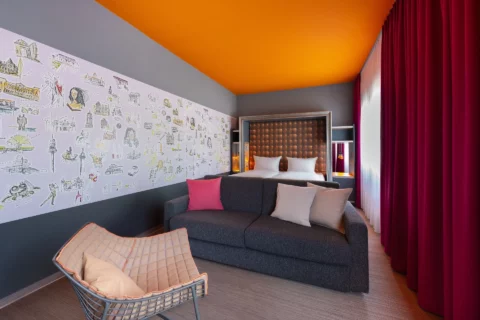 Junior-Suite – Perspektive vom Wohnbereich zum Bett. Designersessel, Sofa mit Kissen, Doppelbett mit gerahmtem Kopfteil, Hannover-Motivwand links, Fenster rechts.